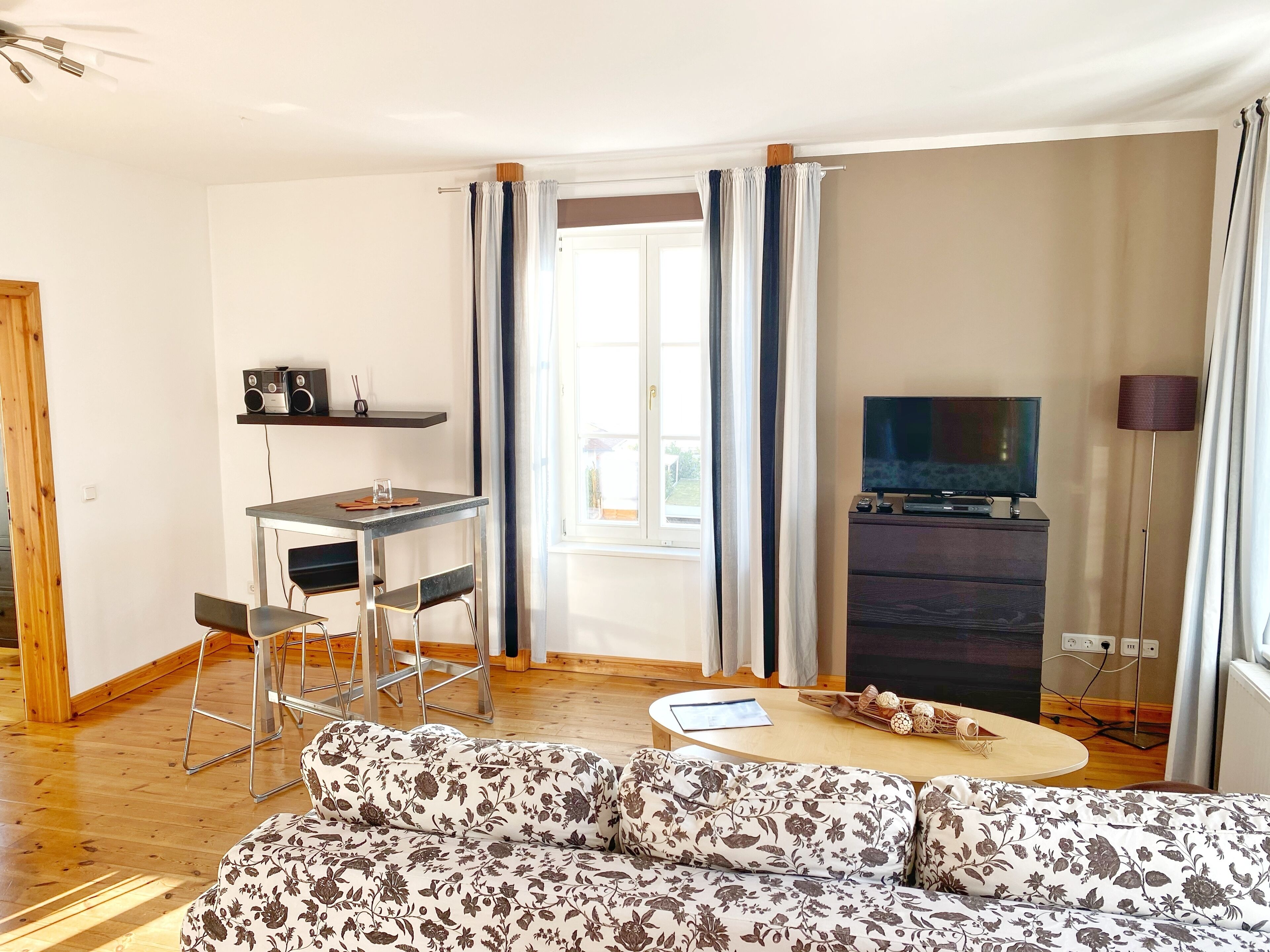 Uma sala de estar com sofá, mesa de jantar com cadeiras, TV em um suporte e janela com cortinas.