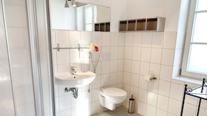 Une salle de bain avec un lavabo blanc, des toilettes et une petite étagère au-dessus des toilettes. Une fenêtre laisse entrer la lumière naturelle.
