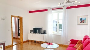 Un salon avec un canapé rouge, un meuble TV blanc, une table blanche et une fenêtre avec des rideaux.