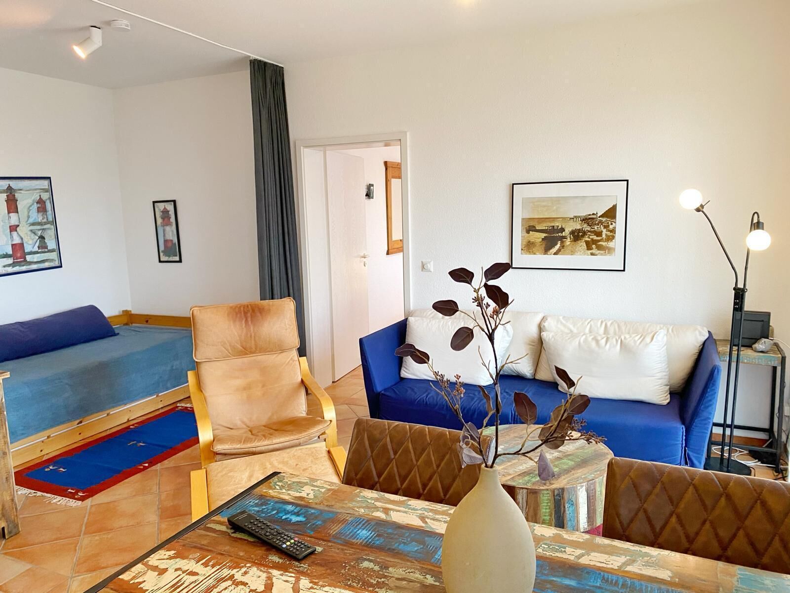 A living room with a sofa, armchairs, a coffee table, and a lamp. There are paintings on the walls and a door in the background.