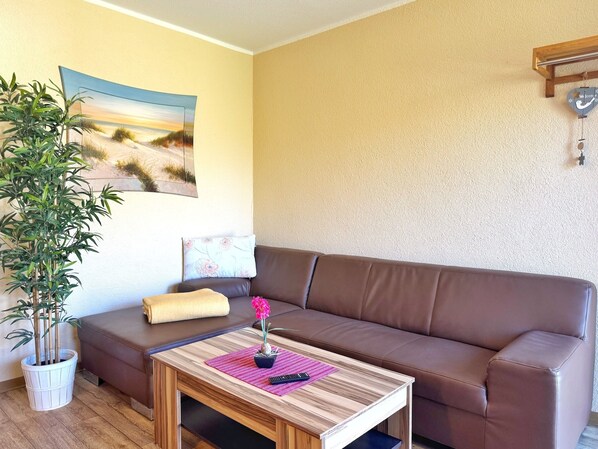 A living room with a brown leather sofa, a wooden coffee table with a remote control, a potted plant, and a painting of a beach scene.