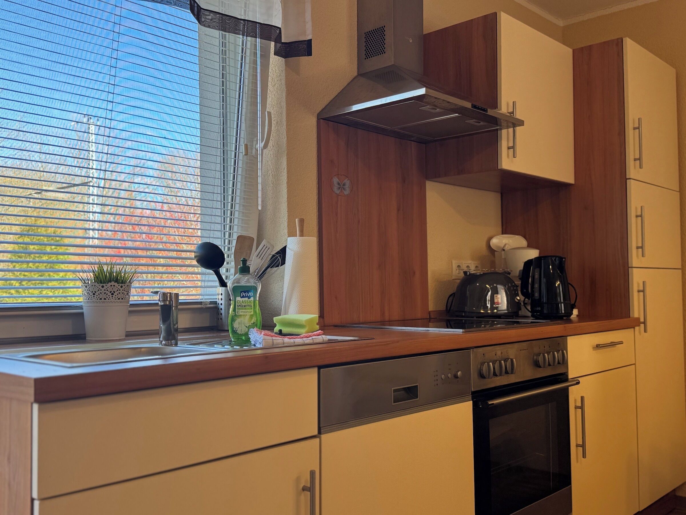 A modern kitchen with wooden cabinets, a stainless steel range hood, a coffee maker, and a window with blinds.