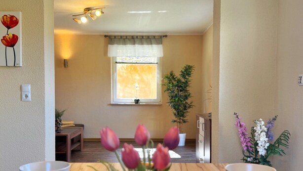 Ein Essbereich mit zwei weißen Schüsseln auf dunklen Untersetzern, einer Vase mit rosa Tulpen, einem Fenster mit Vorhang und einer Topfpflanze.
