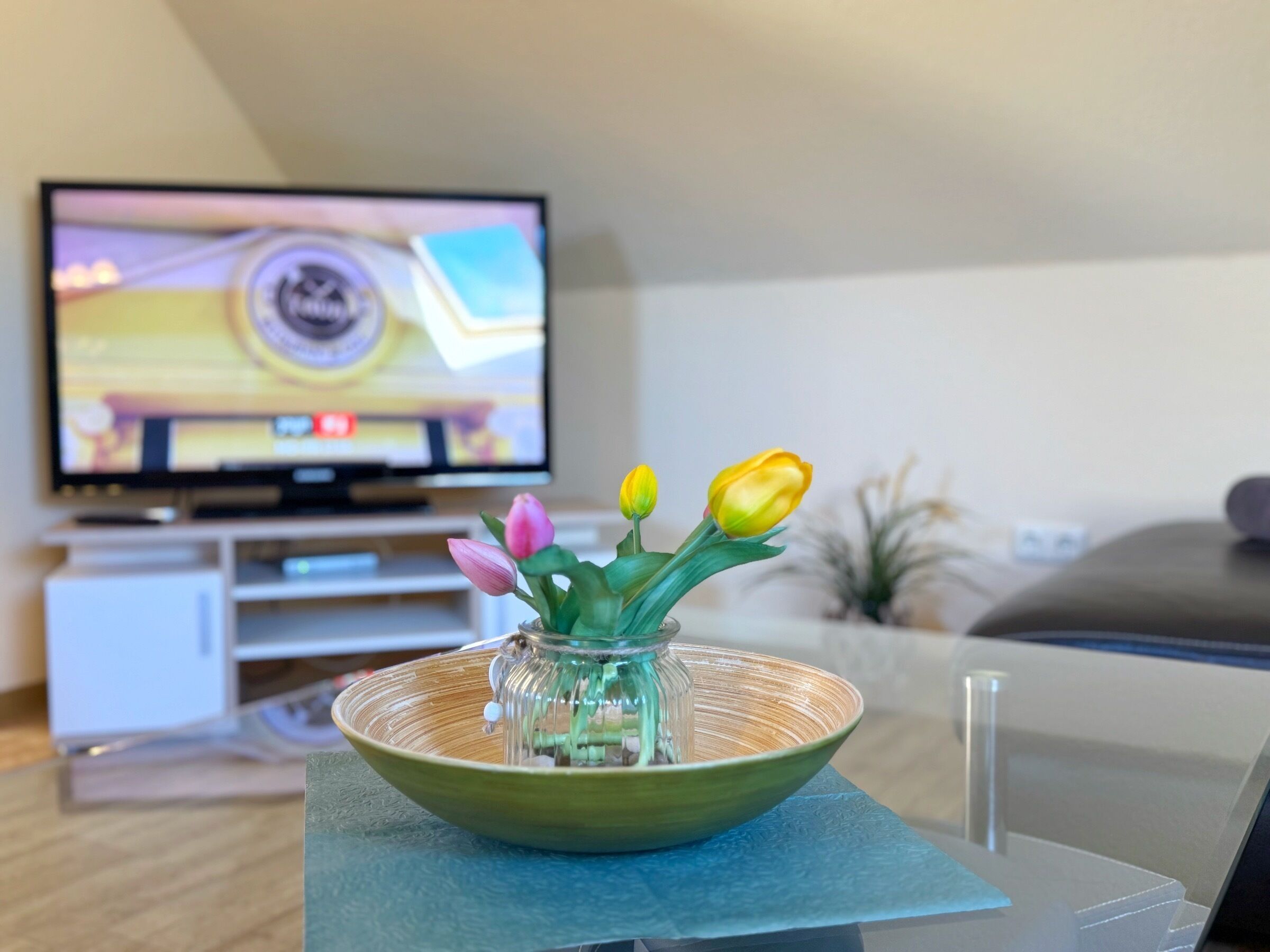 Ein Wohnzimmer mit Fernseher, der ein Programm zeigt, einer Tischvase mit Blumen und einer Couch im Hintergrund.