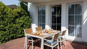 A patio table with four chairs, a small table with food and drinks, and a hedge in the background. - Vacation apartment Neddesitz No. 51 (Neddesitz)