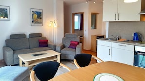 A modern living room with a grey sofa, a small table, and a dining table with chairs. There is a kitchen area with white cabinets and a sink. - Ferienwohnung 2, Am Hochzeitsberg 3A (Breege)