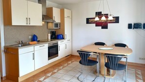A modern kitchen with white cabinets, a stove, and a microwave. There is a dining table with two chairs and a hanging light fixture above it. - Ferienwohnung 2, Am Hochzeitsberg 3A (Breege)