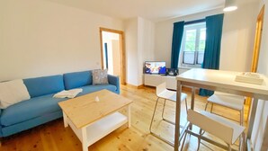 A modern living room with a blue sofa, a wooden coffee table, a white dining table with chairs, and a TV.