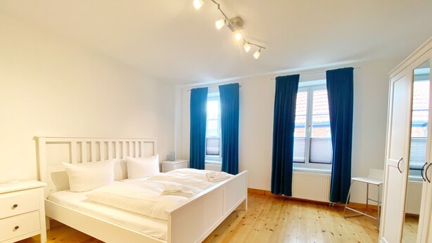 A bedroom with a white bed, wooden flooring, a nightstand, a window with blue curtains, and a radiator.