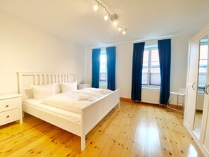 A bedroom with a white bed, wooden flooring, a nightstand, a window with blue curtains, and a radiator.