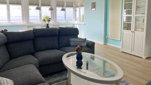 A modern living room with a grey sectional sofa, a glass coffee table, and a view of the sea through large windows. - Villa Anna - App. 41 (Sassnitz)