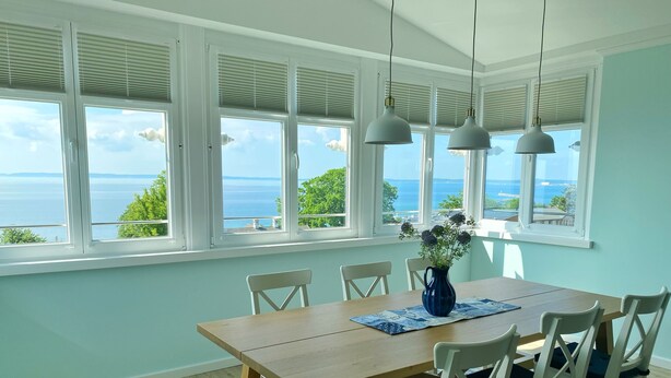 A dining area with a wooden table, white chairs, a vase with flowers, and a view of the sea through large windows with blinds. - Villa Anna - App. 41 (Sassnitz)