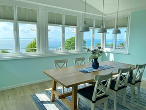 Ein Essbereich mit Holztisch, weißen Stühlen, einer Blumenvase und Meerblick durch große Fenster mit Jalousien.