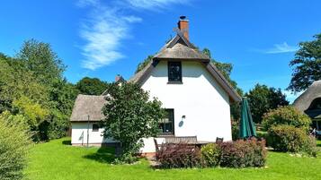 A white cottage with a thatched roof, surrounded by lush greenery and a clear blue sky.
