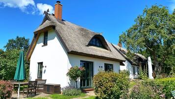 A traditional thatched-roof house with a white exterior, a wooden bench, and a green umbrella in the garden.