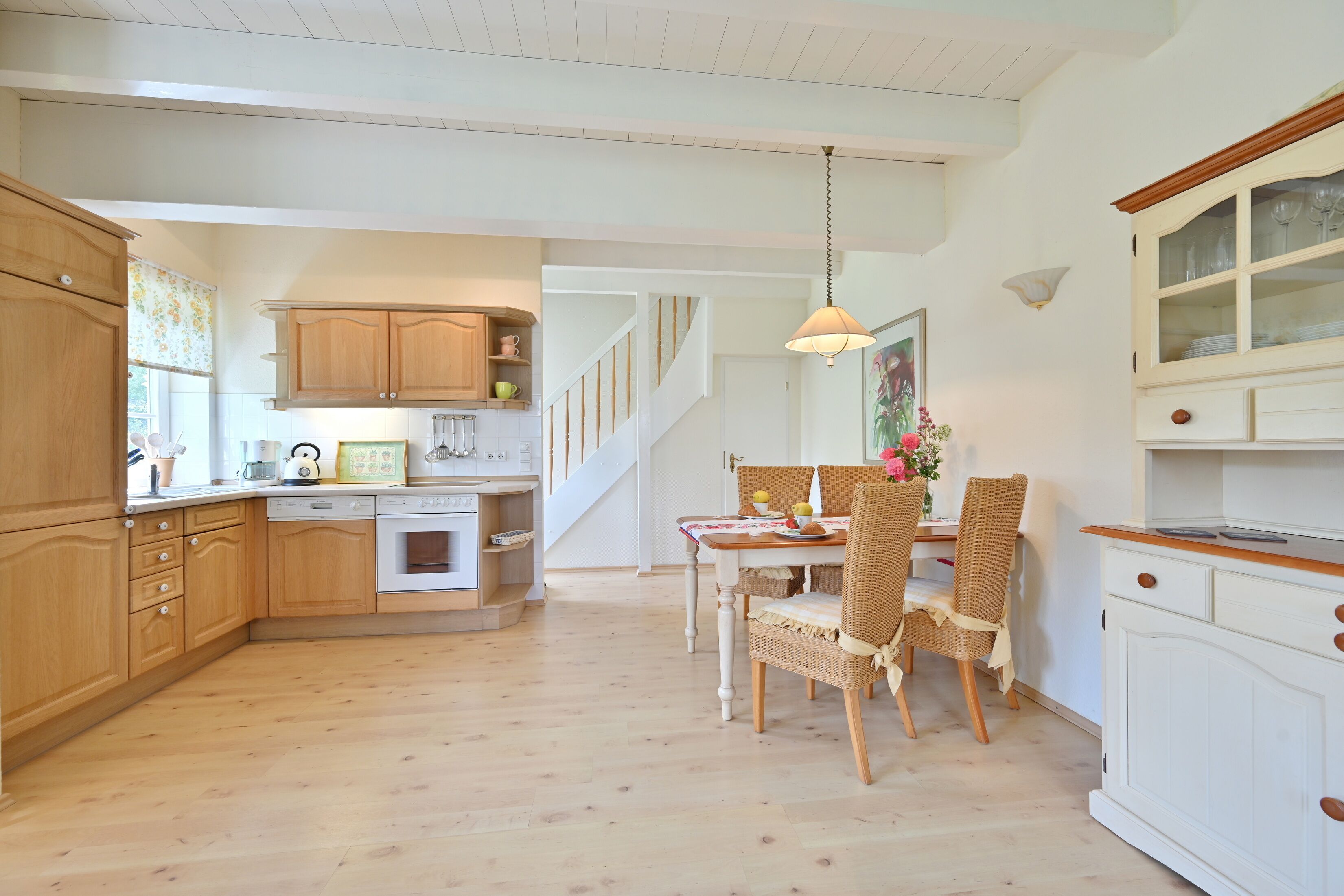 Une cuisine avec des meubles en bois, un coin repas avec des chaises en osier et un escalier menant à un autre niveau.