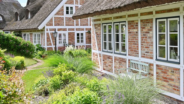 Een traditioneel bakstenen huis met rieten dak, witte raamkozijnen en een tuin met verschillende planten.