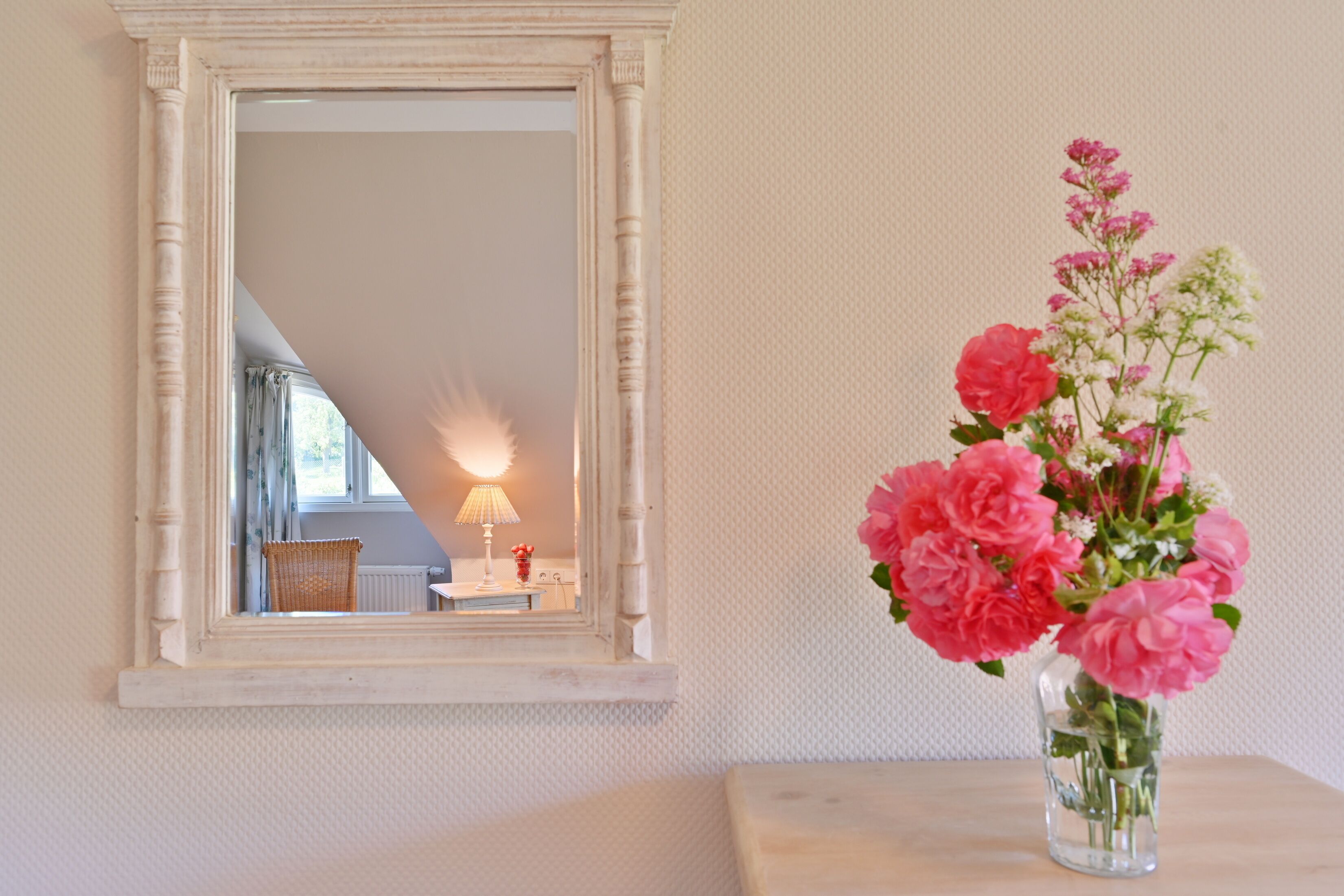 Un miroir avec un cadre blanc, un vase rempli de fleurs roses et une lampe posée sur une table.
