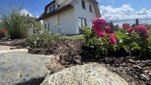 A white house with a thatched roof, surrounded by a garden with pink flowers and a wooden fence. - Ferienhaus "rugard" mit Panoramasauna und Boddenblick (Dranske)