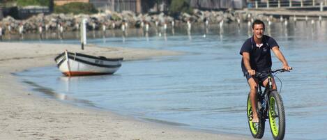 A person riding a bicycle on a sandy beach with calm water and a boat in the background.