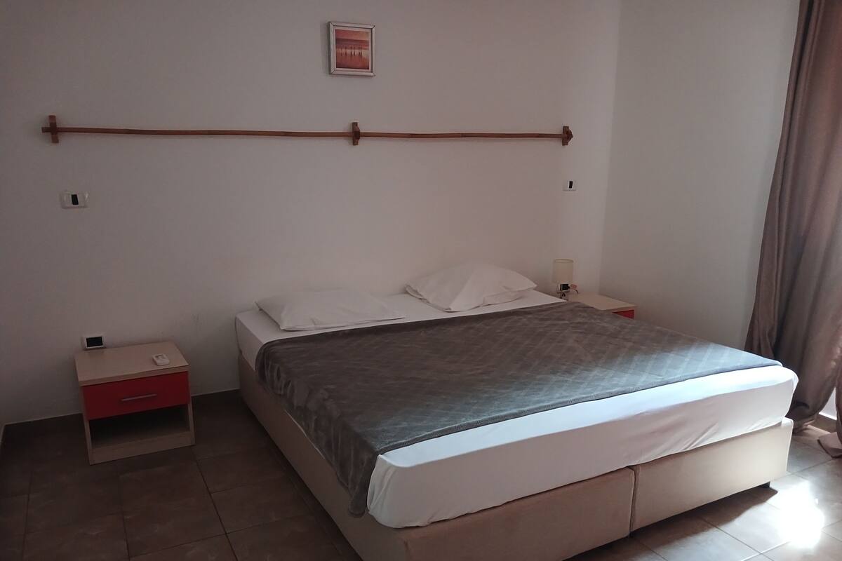 A bedroom with a bed, a nightstand, a wall-mounted shelf, and a window with curtains.