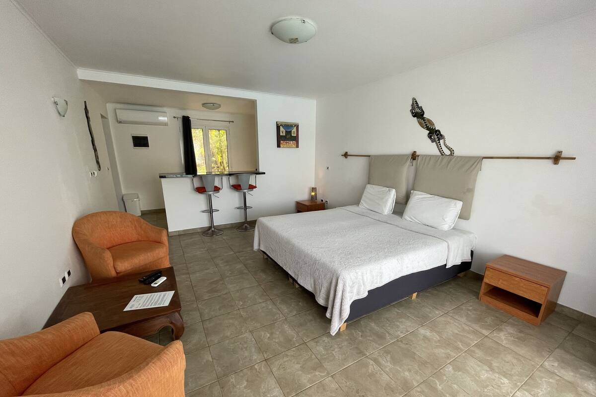 A hotel room with a bed, a chair, a small table, a nightstand, and a kitchenette area with a bar stool.