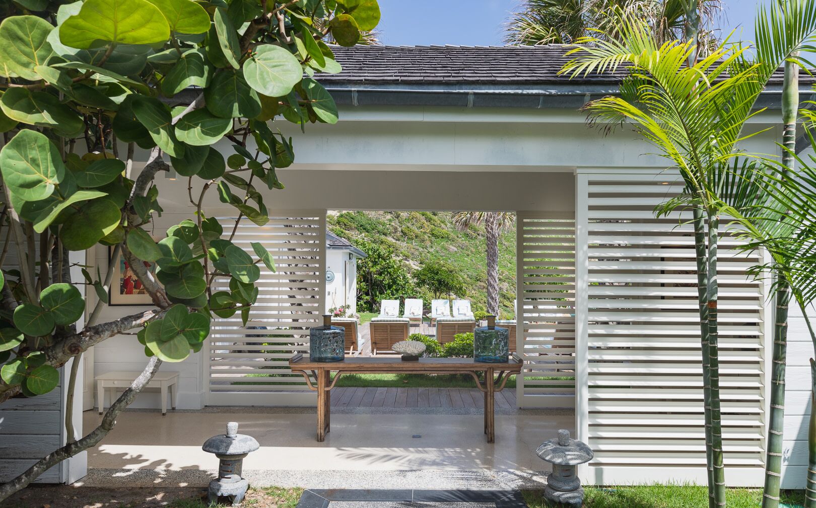 A covered outdoor seating area with a table and chairs, surrounded by greenery and a white fence.
