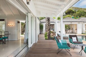 Un espacio al aire libre techado con suelo de madera, una mesa de comedor con sillas, y una zona de estar junto a la piscina con sillones.