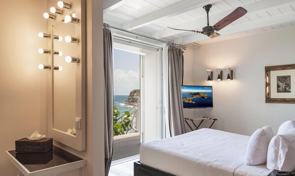 Une chambre d’hôtel avec un grand lit, une télévision, un ventilateur de plafond, un miroir avec éclairage intégré et une vue sur l’océan.