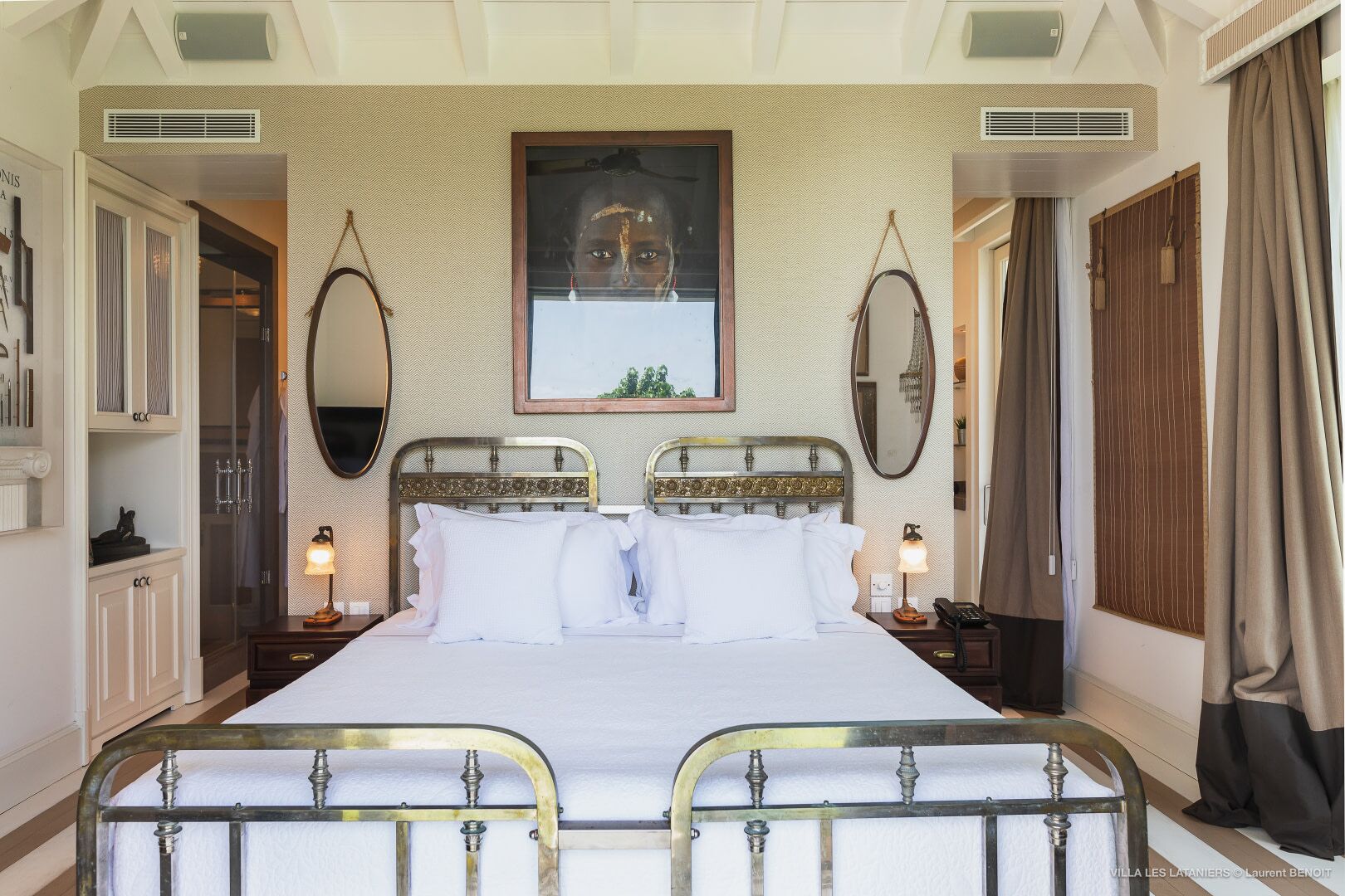 A double bed with a metal frame, two bedside tables with lamps, a large mirror above the bed, and a patterned wallpaper.