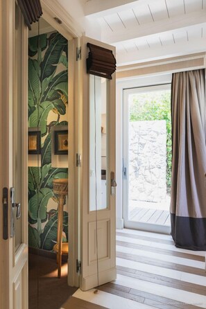Un couloir avec une porte donnant sur un balcon, un plancher en bois et un mur orné d’un motif de feuilles tropicales.