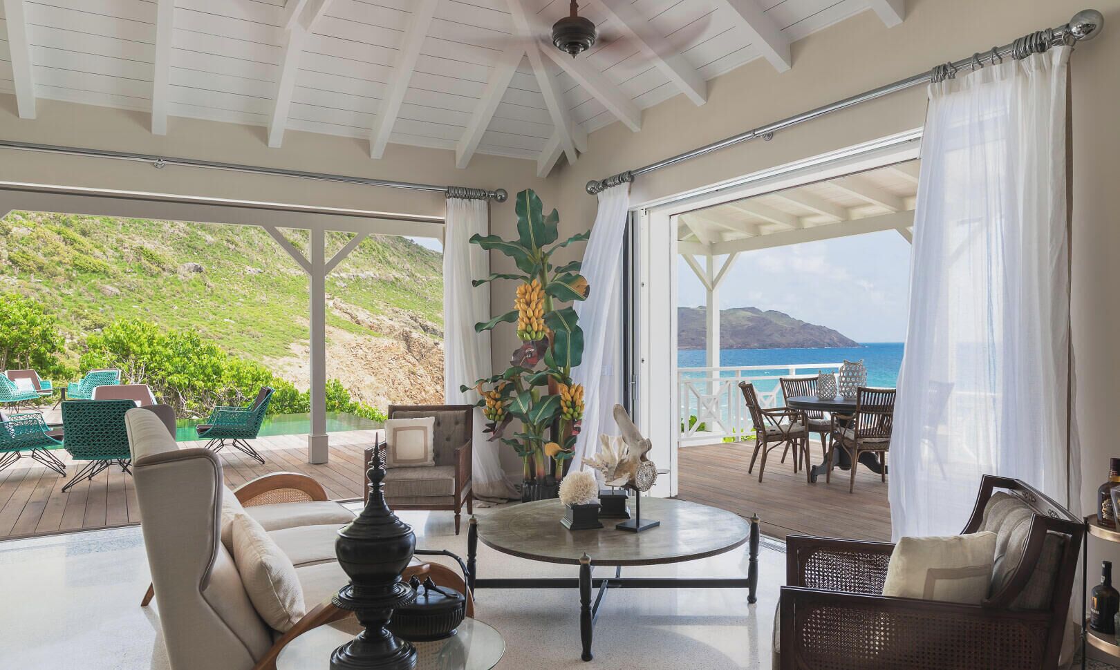 Amplio espacio interior con vista al océano, amoblado con sillas y una mesa, y decorado con plantas y una lámpara.