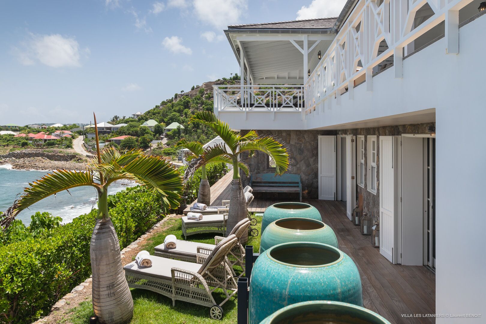 Une villa en bord de mer dotée d’un balcon donnant sur la mer, de chaises longues et de grands pots décoratifs.