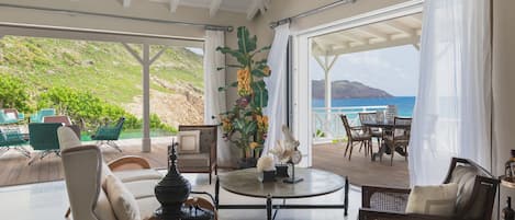 A spacious indoor area with a view of the ocean, furnished with chairs and a table, and decorated with plants and a lamp.