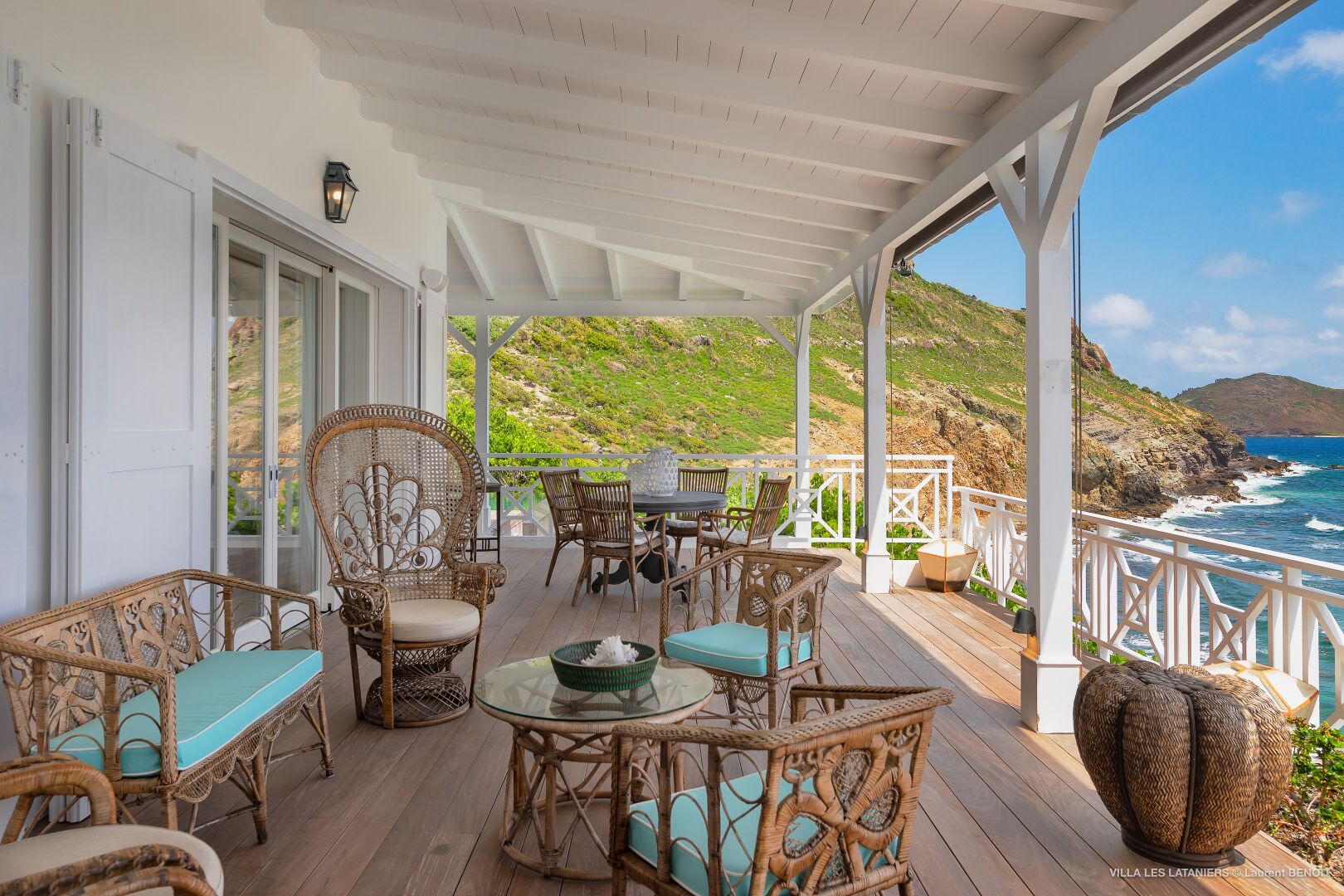 Un espace extérieur abrité, aménagé avec des meubles en osier et une table en verre, offrant une vue sur un paysage côtier.