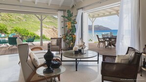 Amplio espacio interior con vista al océano, amoblado con sillas y una mesa, y decorado con plantas y una lámpara.