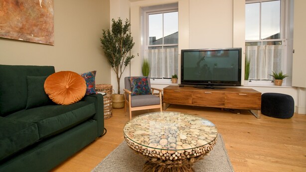 Ein modernes Wohnzimmer mit einem grünen Sofa, einem runden Glastisch, einer Holz-TV-Konsole und einem großen Fenster mit Vorhängen.