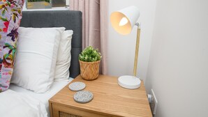 Ein ordentlich bezogenes Bett mit weißer Bettwäsche, einem grauen Kopfteil und einem farbenfrohen Blumenkissen. Am Bett steht ein kleiner Nachttisch mit einem gewebten Korb, der grüne Pflanzen und zwei dekorative Teller enthält. Eine weiße Lampe mit orangem Lampenschirm steht auf dem Nachttisch.
