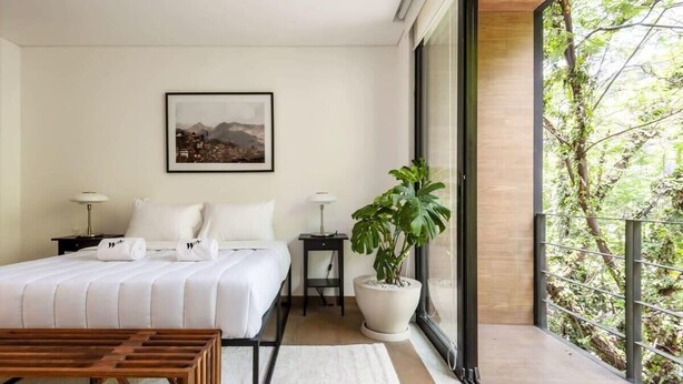A modern hotel room with a large bed, bedside tables, a potted plant, and a balcony with a view of greenery. - w* | Inspiring 3BR Duplex with Terrace in Polanco (Ciudad de México)