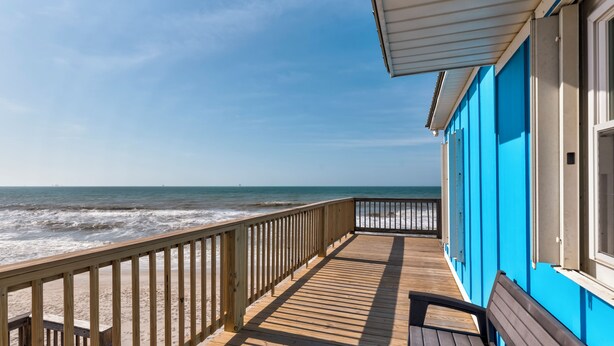 A wooden deck with a railing overlooking the ocean, a bench, and a building with blue walls. - Blue Sky Breeze - Gulf Front, Pet Friendly on the beach (Dauphin Island)