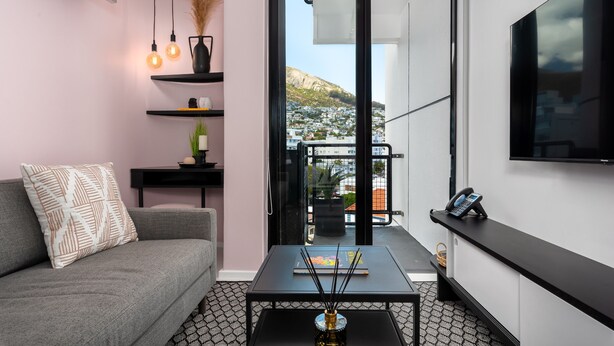 A modern living room with a sofa, coffee table, and shelves. A balcony with a view of a city and mountains is visible through an open door. - Stylish Living at The Flamingo! (Cape Town)