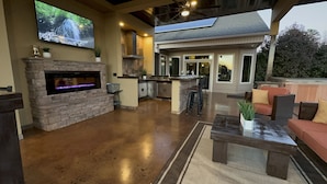 A spacious outdoor living area with a stone fireplace, a flat-screen TV mounted on the wall, a kitchen area with a bar, and outdoor seating. - 50% OFF - No Stairs (Sevierville)