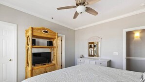 A bedroom with a bed, a television on a wicker stand, a ceiling fan, a dresser, and a mirror.