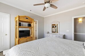A bedroom with a bed, a television on a wicker stand, a ceiling fan, a dresser, and a mirror.
