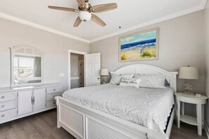 Una camera da letto con un letto, una ventola a soffitto, un quadro raffigurante una spiaggia, una cassettiera e un comodino con una lampada.