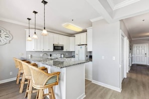 A modern kitchen with white cabinets, a granite countertop, and a bar area with wicker chairs. - Paradise Villa 4B/4BA Breathtaking Ocean View (Ocean Isle Beach)