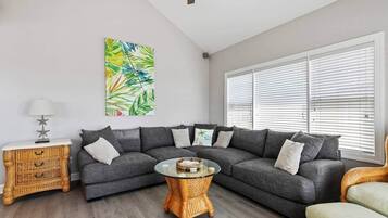 Un salon avec un canapé d’angle gris, une table basse ronde, une table d’appoint en osier et une grande fenêtre avec des stores.
