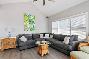 Ein Wohnzimmer mit einem grauen Ecksofa, einem runden Couchtisch, einem Korbtisch und einem großen Fenster mit Jalousien.