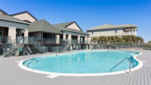A swimming pool with a wooden deck, a covered area with stairs, and buildings in the background.