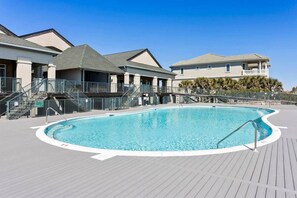 A swimming pool with a wooden deck, a covered area with stairs, and buildings in the background.